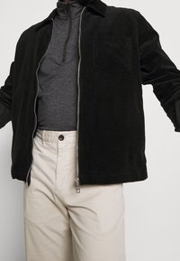 Zwarte corduroy jack met een ritssluiting aan de voorkant, kraag en een borstzak, gedragen over een donkergrijze top en beige broek.