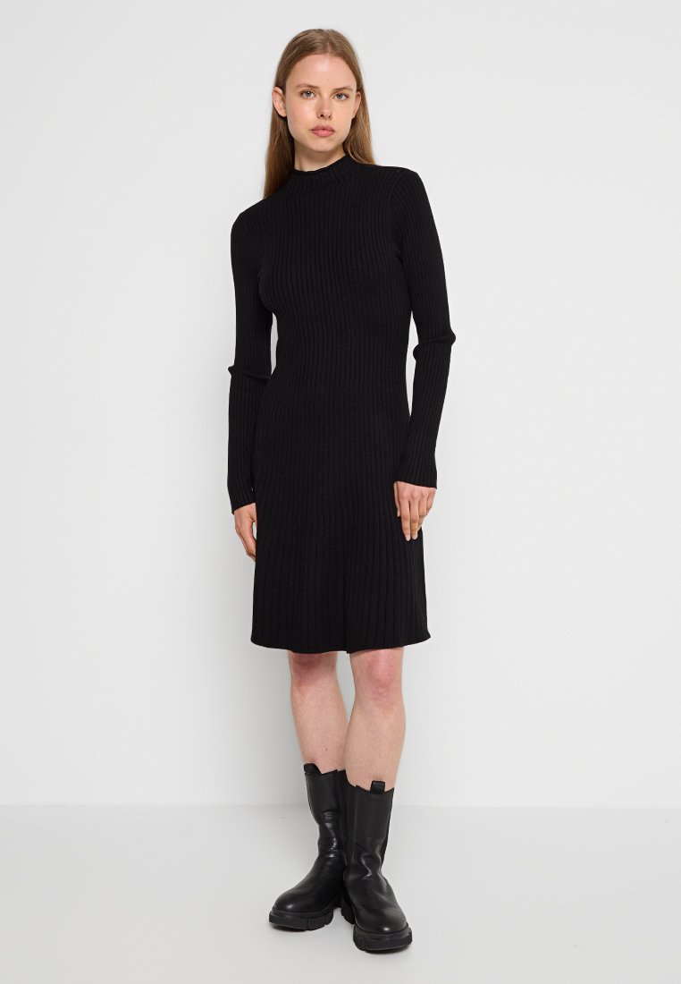 Schwarzes, geripptes Kleid mit hohem Kragen, langen Ärmeln und figurbetonter Silhouette. Kombiniert mit klobigen, schwarzen Stiefeletten. Einfacher, strukturierter Stoff.