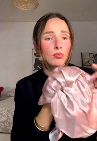 Frau mit braunen Haaren hält und begutachtet eine glänzende pinke Satinmütze in einem sanft beleuchteten Schlafzimmer mit Wandkunst und einer Hängeleuchte.