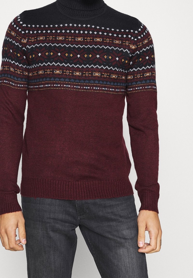 Mann mit einem bordeauxfarbenen Pullover mit mehrfarbigen nordischen Mustern über der Brust und den Schultern, kombiniert mit dunkelgrauen Jeans.