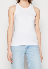 Bílý žebrovaný top bez rukávů s kulatým výstřihem, v kombinaci se světle modrými džíny.