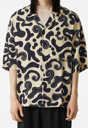 Chemise beige à manches courtes avec boutons, ornée de grandes formes abstraites noires et de points blancs, portée par une personne portant un pantalon noir.