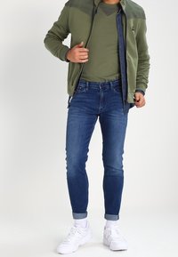 Man wearing green jacket, olive shirt, blue rolled-up jeans, white socks, and white sneakers, standing against plain background.