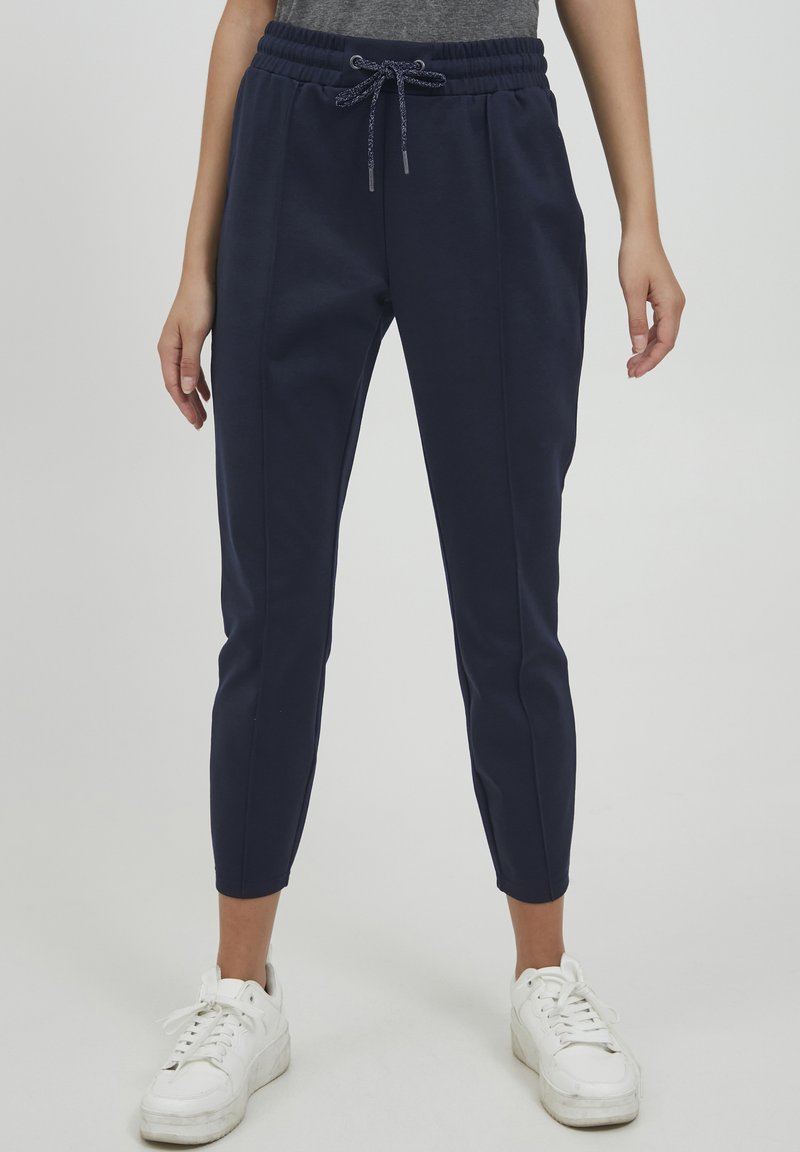 Navy Jogginghosen mit elastischem Bund und Kordelzug. Weicher, glatter Stoff mit schmal zulaufenden Beinen. Das Modell trägt weiße Sneakers.