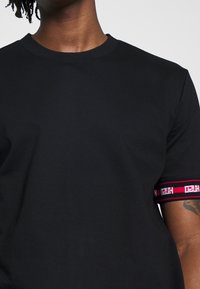 Camiseta negra de manga corta con cuello redondo. Presenta una banda a rayas rojas y blancas en la manga con texto repetido. Textura suave.