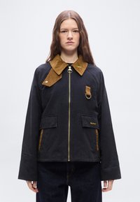 Νεαρή γυναίκα που φοράει μαύρο μπουφάν με καφέ γιακά και λεπτομέρειες, χρυσό φερμουάρ και εμπρόσθιες τσέπες, στέκεται μπροστά σε απλό ανοιχτό φόντο.