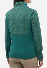 Femme portant une veste zippée en teal et vert chiné vue de dos, associée à un pantalon beige.