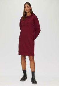 Burgunderfarbendes Kapuzenkleid mit seitlichen Schlitzen, aus weichem Material gefertigt. Getragen mit schwarzen Knöchelstiefeln, hat einen lockeren Schnitt und eine verstellbare Kapuze.