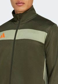 Green athletic jacket with a high collar, featuring a light green stripe across the chest and orange logo accents. Zippered front.