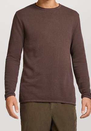 Homme portant un pull marron ajusté à manches longues et texturé, avec un pantalon vert olive, debout devant un fond clair uni.