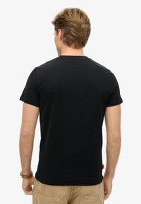 Sort bomulds T-shirt med rund halsudskæring, korte ærmer og lige kant. Har et rødt mærke på den nederste venstre side.