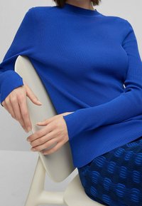 Pull bleu côtelé à col montant et manches longues, associé à une jupe bleue à motifs texturés. Position assise avec les mains posées.