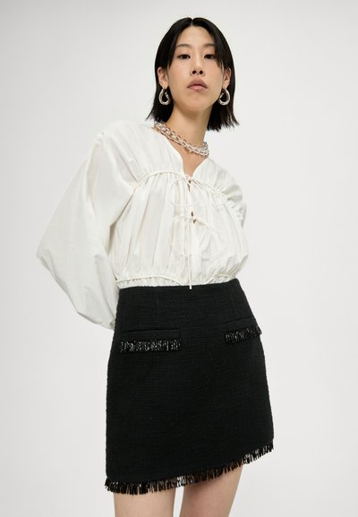 Blouse blanche avec des détails froncés et un col en V, associée à une mini-jupe noire texturée dotée de poches accentuées de franges.