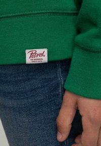 Handen vilar vid fickan på blå denimjeans, iklädd en grön sweatshirt med en etikett som säger "Petrol No Barriers Since 1973".