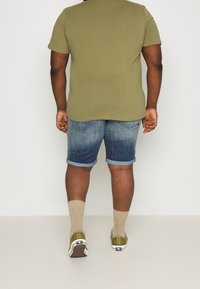 Camiseta verde oliva, pantalones cortos de mezclilla con los puños enrollados, calcetines beige y zapatos verdes. El conjunto presenta un ajuste casual y relajado.