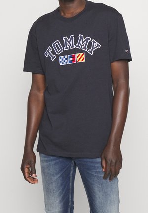 Mężczyzna w czarnym t-shircie Tommy z wzorami morskich flag w granatowym, czerwonym i żółtym kolorze na niebieskich dżinsach, stojący na jednolitym tle.