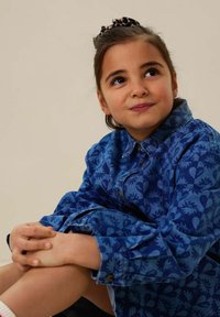 Chemise en denim bleu avec un design à motifs comprenant des cœurs et des motifs floraux. La chemise a un col et des boutons, avec des manches retroussées.