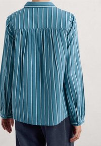 Person wearing a loose teal blouse with vertical white stripes, long sleeves, and pleats on the upper back, paired with dark ribbed pants.