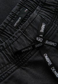 Tejido de mezclilla negra con costuras visibles. Las etiquetas muestran "Ombre" y "Denim" en texto blanco. Presenta costuras con volantes y un ajuste estándar.