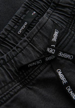 Tessuto in denim nero con cuciture visibili. Le etichette mostrano "Ombre" e "Denim" in testo bianco. Presenta cuciture arricciate e una vestibilità standard.