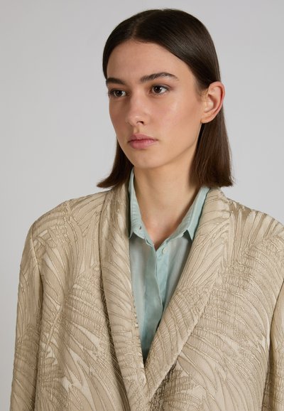 Giovane donna con capelli castani lisci che indossa una giacca beige strutturata sopra una camicia celeste con colletto, che guarda leggermente di lato.