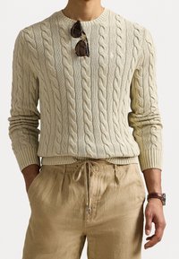 Pull en tricot torsadé crème avec des poignets et un ourlet côtelés, associé à un pantalon en lin beige. Des lunettes de soleil reposent sur le décolleté.