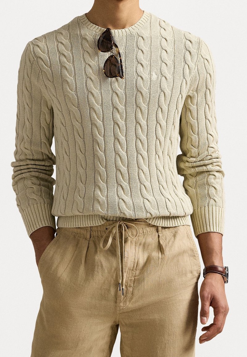 Pull en tricot torsadé crème avec des poignets et un ourlet côtelés, associé à un pantalon en lin beige. Des lunettes de soleil reposent sur le décolleté.