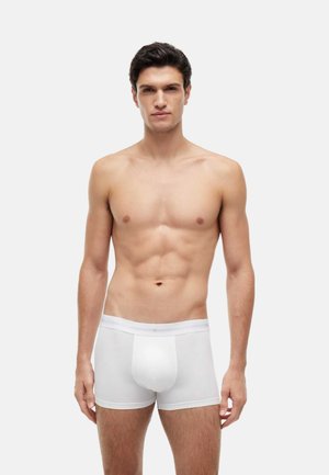 Joven con cabello corto y oscuro, complexión muscular, vestido con calzoncillos tipo bóxer blancos, de pie frente a un fondo liso de color gris claro.