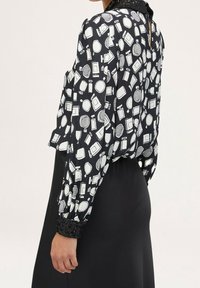 Blusa nera con motivo di prodotti di bellezza bianchi, maniche lunghe e polsini decorati con paillettes. Tessuto leggermente trasparente, vestibilità comoda e apertura a chiave sul retro.