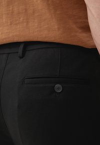 Zwarte broek met een gladde textuur, voorzien van een achterzak en een zwarte knoopaccent. Gepaard met een bruine, gestructureerde top.