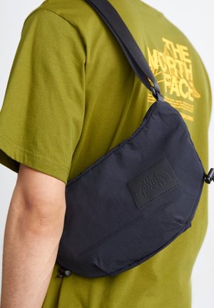 Osoba nosząca oliwkowozieloną koszulkę The North Face z czarnym paskiem i saszetką The North Face przewieszoną przez ramię.