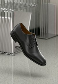 Black leather dress shoe with a polished finish, lace-up design, slightly pointed toe, and a low heel, set against a minimalist background.