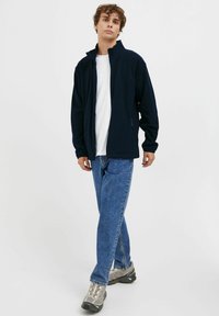 Tumed fleece-jakk, millel on kõrge krae ja tõmblukk, kantud valge T-särgi peal, koos siniste teksade ja hallide tossudega.