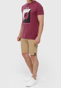T-shirt grafico bordeaux con collo rotondo, abbinato a pantaloni corti beige arrotolati con fibbia laterale e scarpe da ginnastica bianche.