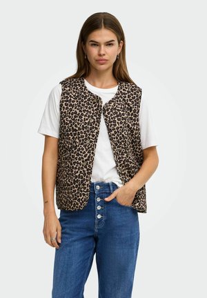 Femme portant un gilet boutonné à imprimé léopard sur un t-shirt blanc et un jean bleu taille haute avec des boutons, debout devant un fond blanc.