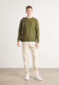Olivgrüner Strickpullover, cremefarbene Hose und weiße Sneakers. Das Model trägt eine grüne Schultertasche und steht vor einem schlichten Hintergrund.