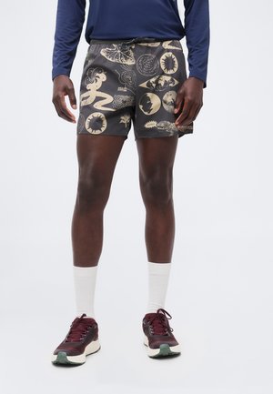 Férfi, fekete rövidnadrággal, bézs absztrakt mintákkal és bordó futócipővel, egyszerű fehér háttér előtt áll.