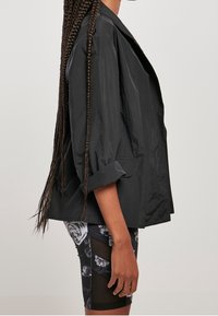 Kvinna med långa flätor iförd en lös svart jacka med uppvikt ärm och svarta shorts med blommönster och meshpaneler, sedd från sidan.