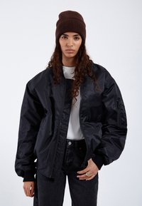 Black bomber jacket with smooth, shiny fabric; ribbed cuffs; paired with a light gray shirt and dark denim. Brown knitted beanie worn.