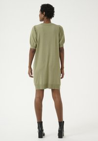 Abito a maglia verde oliva, lungo fino al ginocchio, con maniche a sbuffo e polsini a coste. Presenta un collo rotondo e una texture uniforme. Abbinato a stivaletti neri al collo.