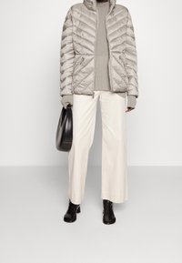Wypełniona, jasnoszara kurtka w wzór chevron, zestawiona z beżowym swetrem w prążki i białymi spodniami o szerokich nogawkach; trzymając czarną torebkę.