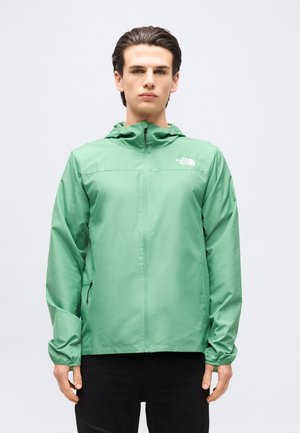 Νεαρός άνδρας φορώντας πράσινο τζάκετ North Face με φερμουάρ και κουκούλα, και μαύρο παντελόνι, στέκεται μπροστά σε απλό ανοιχτόχρωμο φόντο.