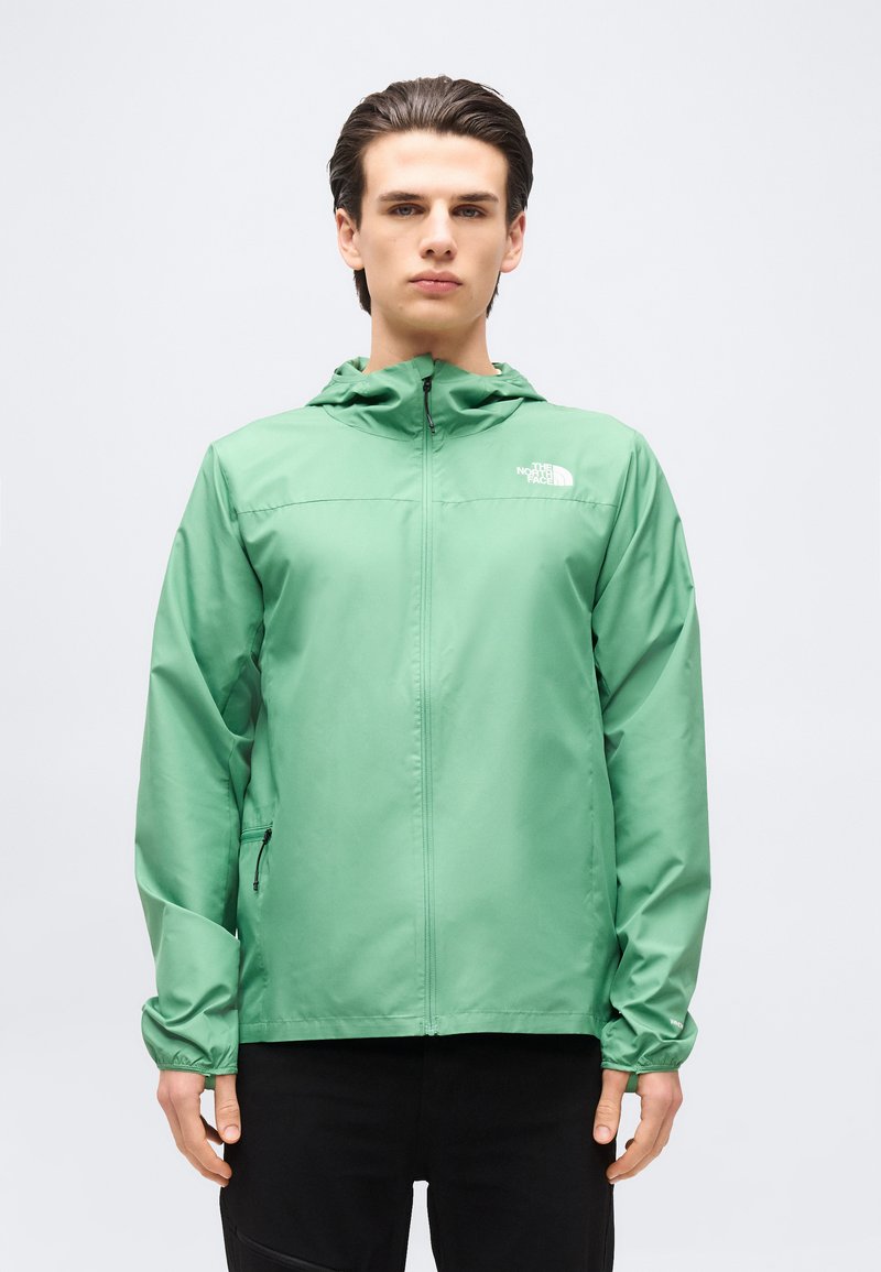 Νεαρός άνδρας φορώντας πράσινο τζάκετ North Face με φερμουάρ και κουκούλα, και μαύρο παντελόνι, στέκεται μπροστά σε απλό ανοιχτόχρωμο φόντο.