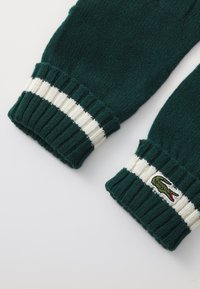 Strikkede grønne hansker med ribbestrikkede mansjetter, som har hvite striper og en liten brodert krokodillelogo på den ene mansjetten. Myk tekstur og tettsittende passform.