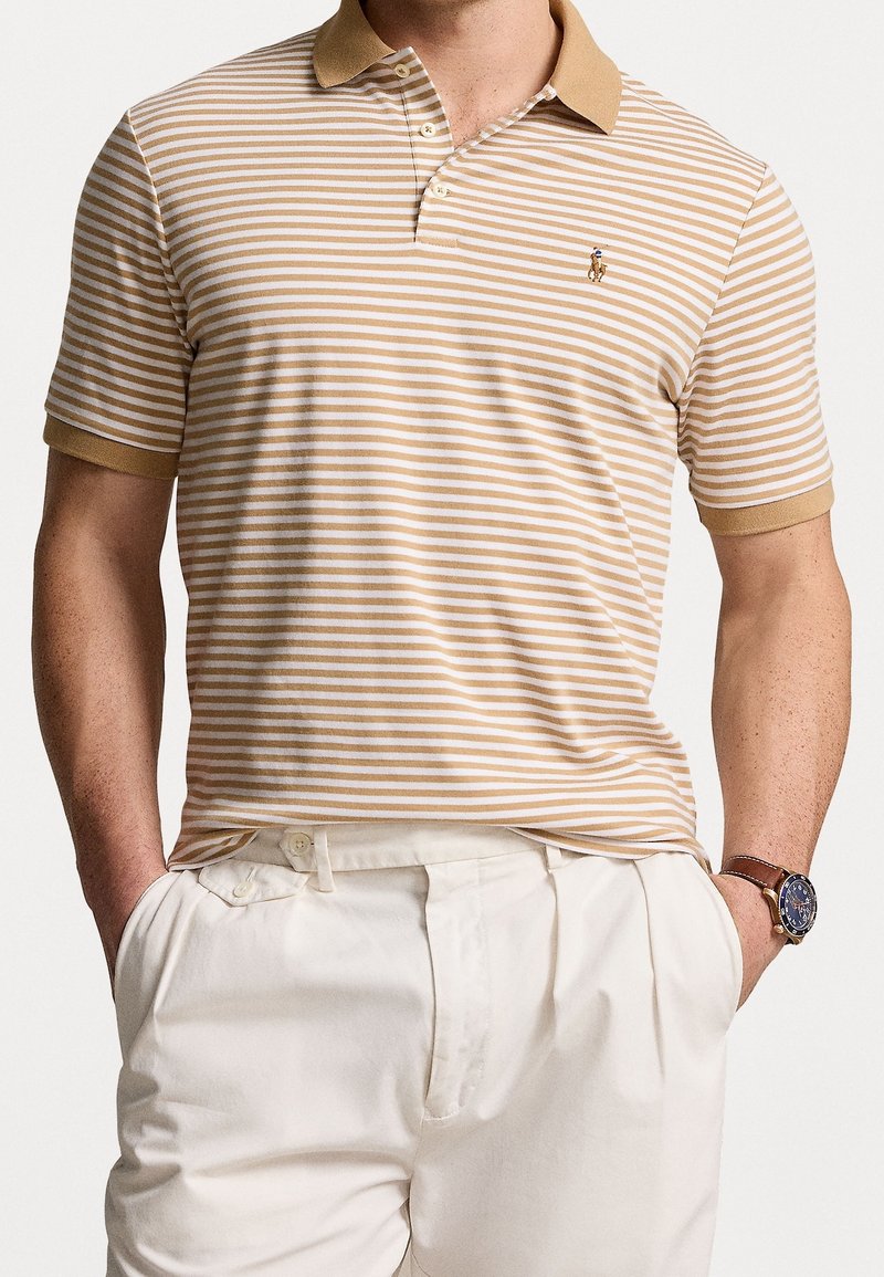 Homme portant un polo à manches courtes rayé beige et blanc, rentré dans un pantalon blanc taille haute, les mains dans les poches et une montre-bracelet.