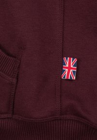Felpa borgogna con una texture morbida, polsini a costine e una piccola toppa con la bandiera britannica sul davanti.