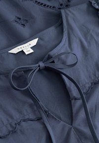 Μπλούζα σε ναυτικό μπλε με κλείσιμο με φιόγκο, κυματοειδείς άκρες και περίτεχνο σχέδιο με κοψίματα. Κατασκευασμένη από ελαφρύ ύφασμα. Ετικέτα μάρκας: Noella.