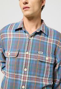 Camisa de cuadros con botones en colores azul, rojo, verde y blanco, hecha de una tela de textura suave, con dos bolsillos en el pecho y etiqueta de marca.
