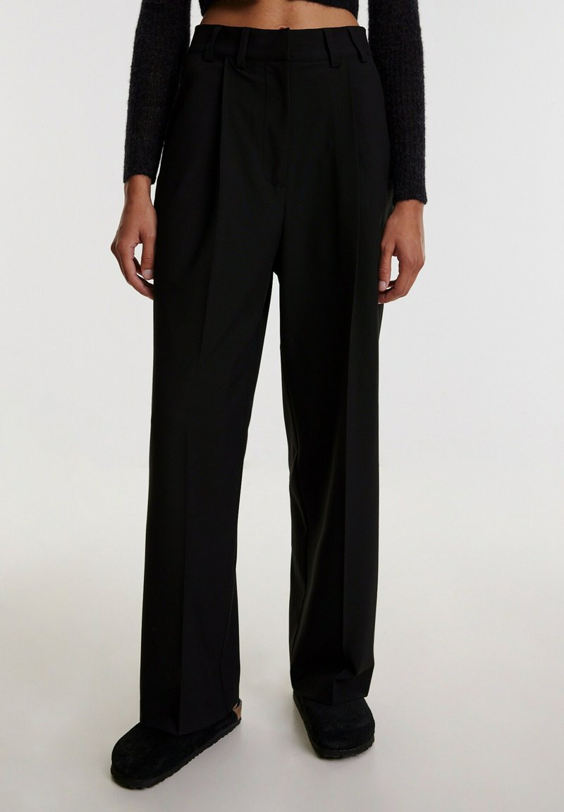 Schwarze, hoch taillierte Hose mit Falten, die eine glatte Textur und ein weites Bein-Design aufweist. Getragen mit schwarzen, weich strukturierten Schuhen.