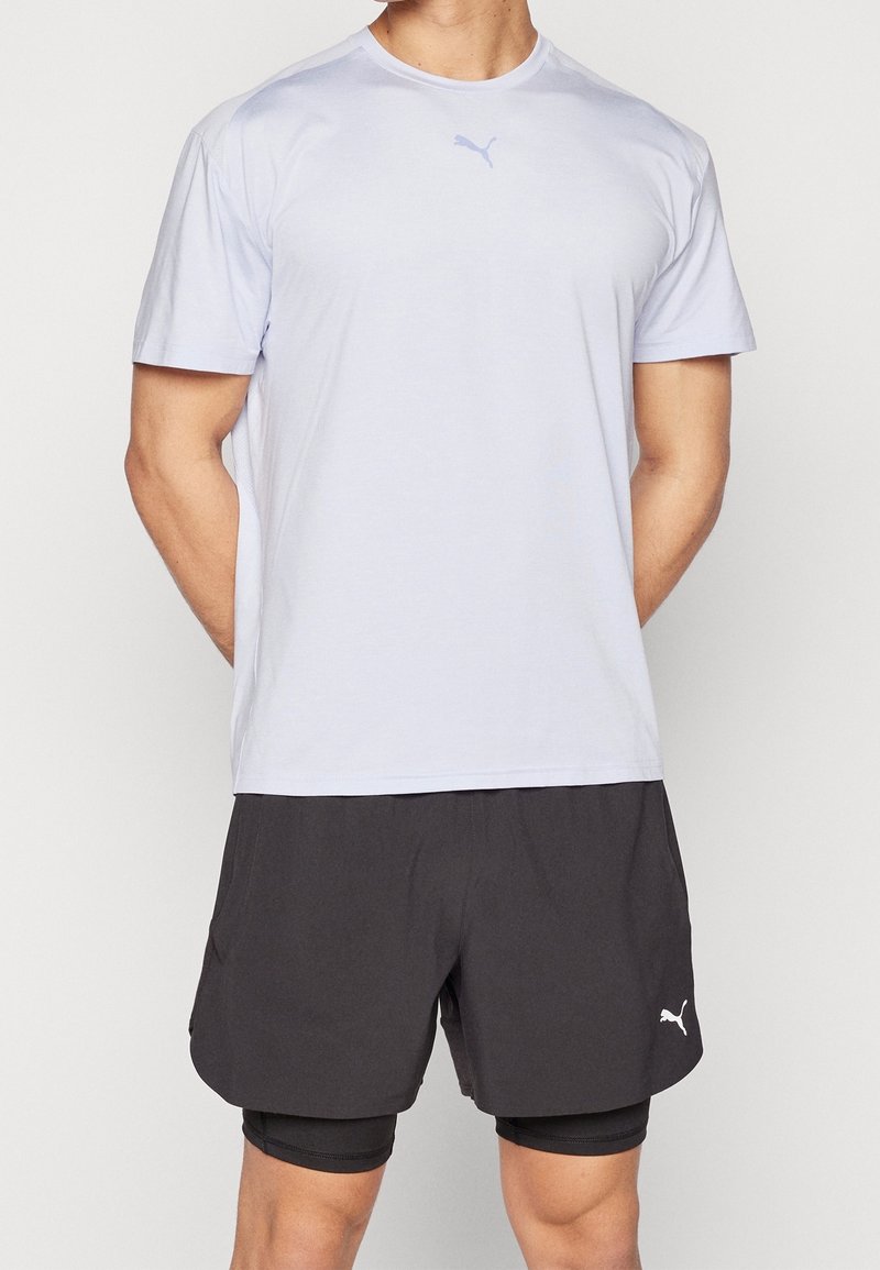 Hombre con una camiseta blanca de manga corta de Puma y pantalones deportivos negros en capas de Puma, con las manos detrás de la espalda, de pie contra un fondo liso.